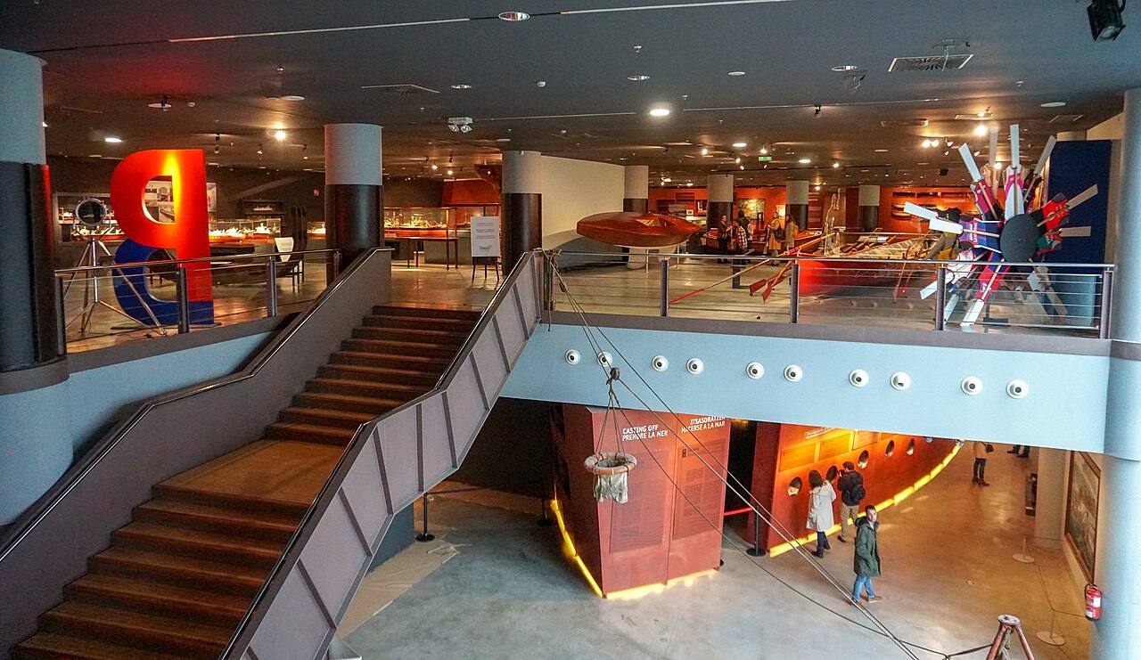 Interior de un museo con exposiciones, escaleras y visitantes